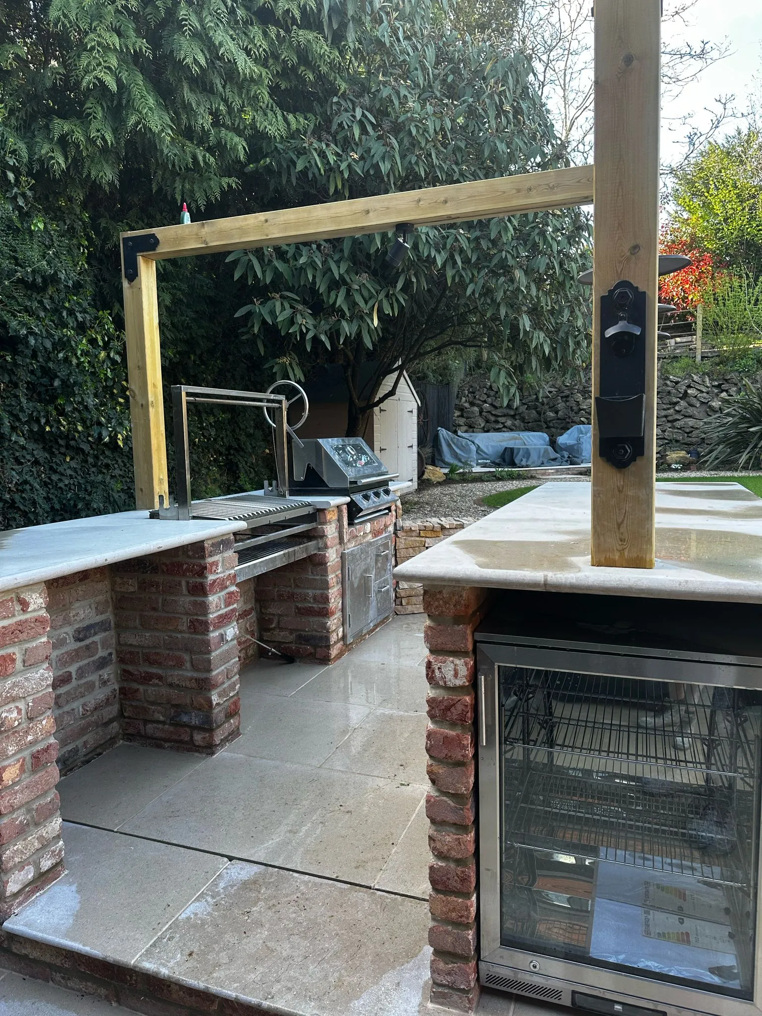 An outdoor fridge built in to reclaimed bricks in the foreground, with an outdoor kitchen worktop, grill, BBQ and storage cupboard also visible